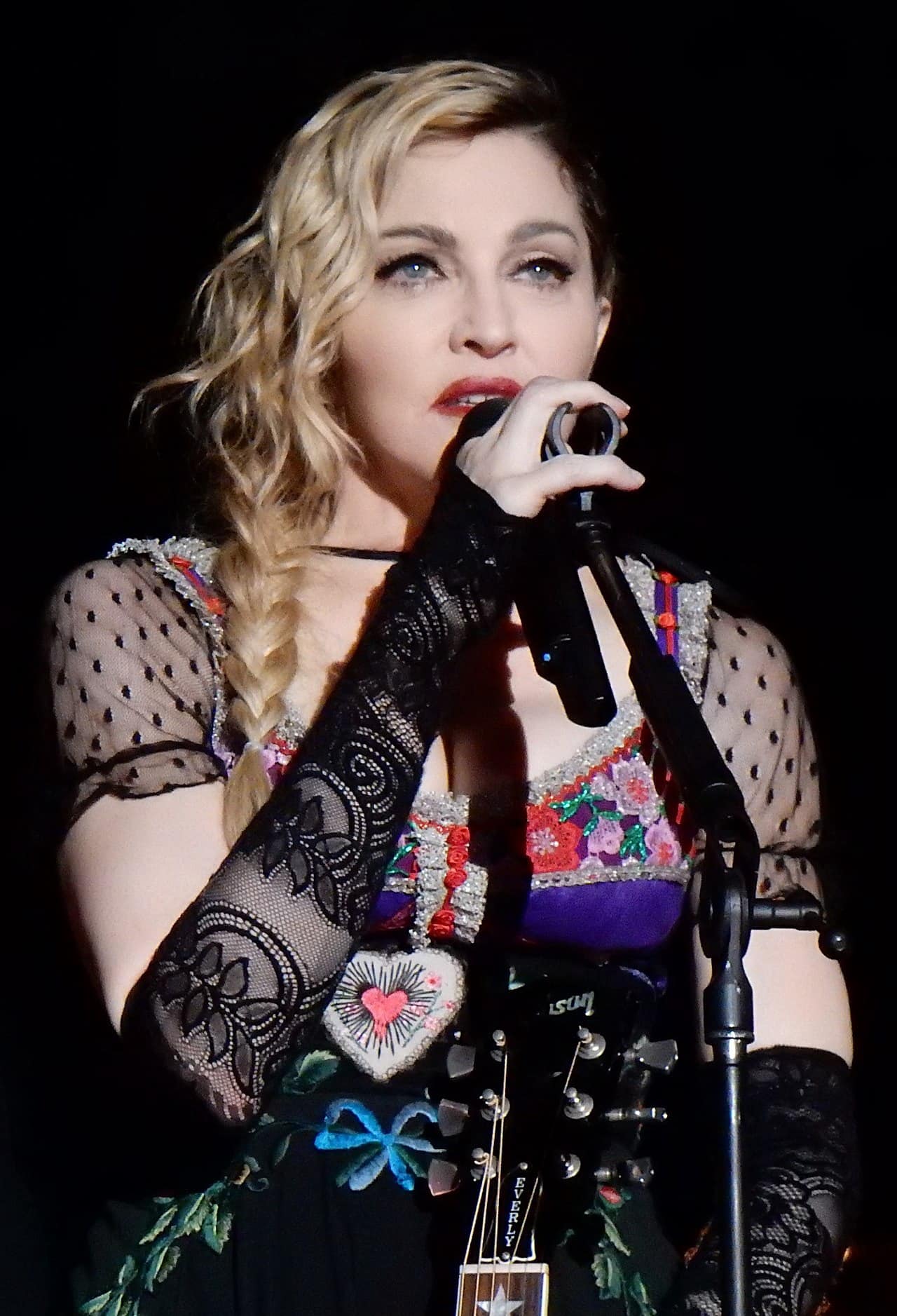 Madonna auf ihrer Rebel Heart Tour in Stockholm (2015) mit schwarz-buntem Kleid und Netzarmstulpen, langer, blonder geflochtener Zopf zur rechten Seite mit Standmikro singend.