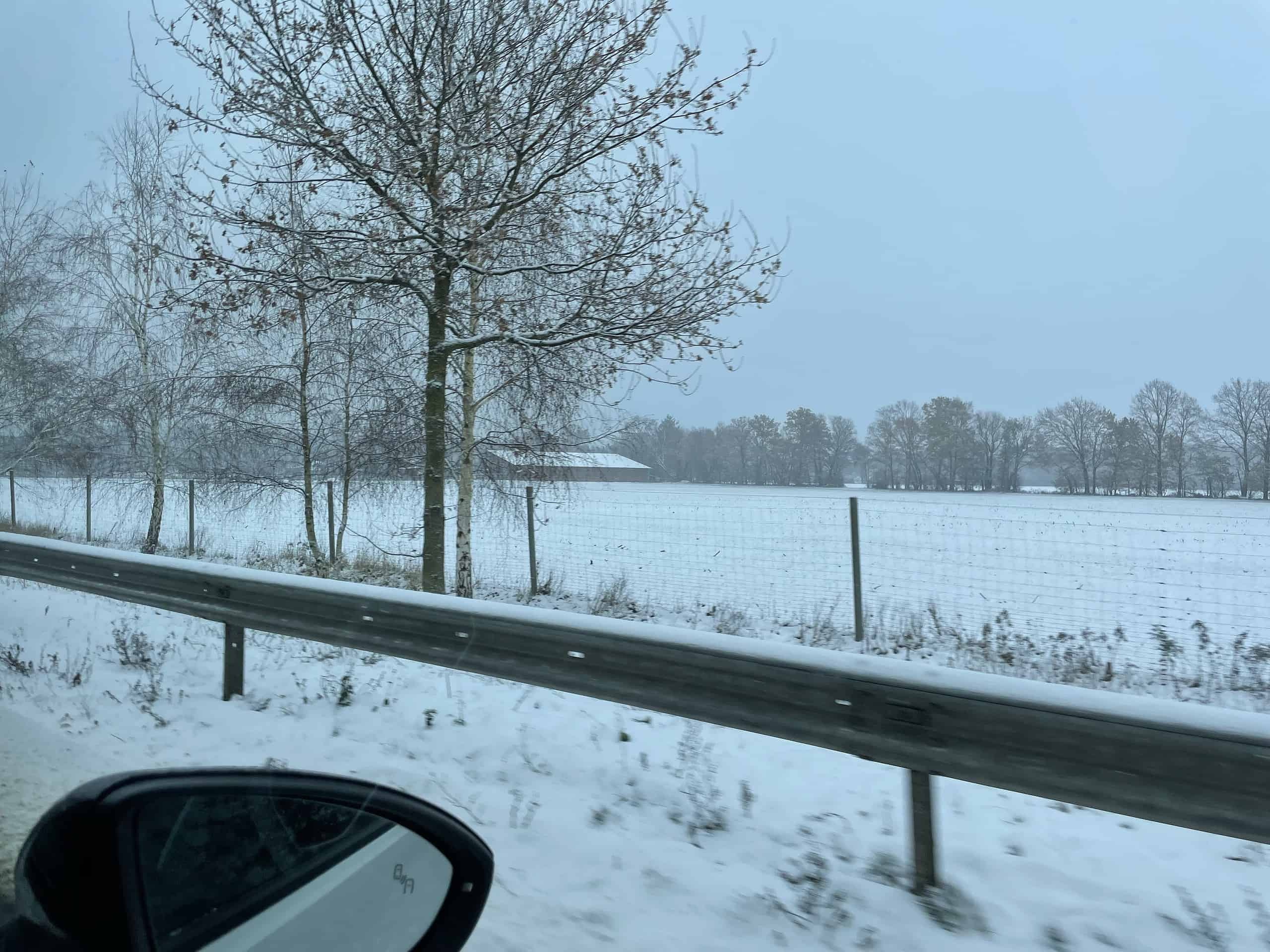 Schneelandschaft rechts vom fahrenden Auto mit Leitplanke und Außenspiegel im Vordergrung.