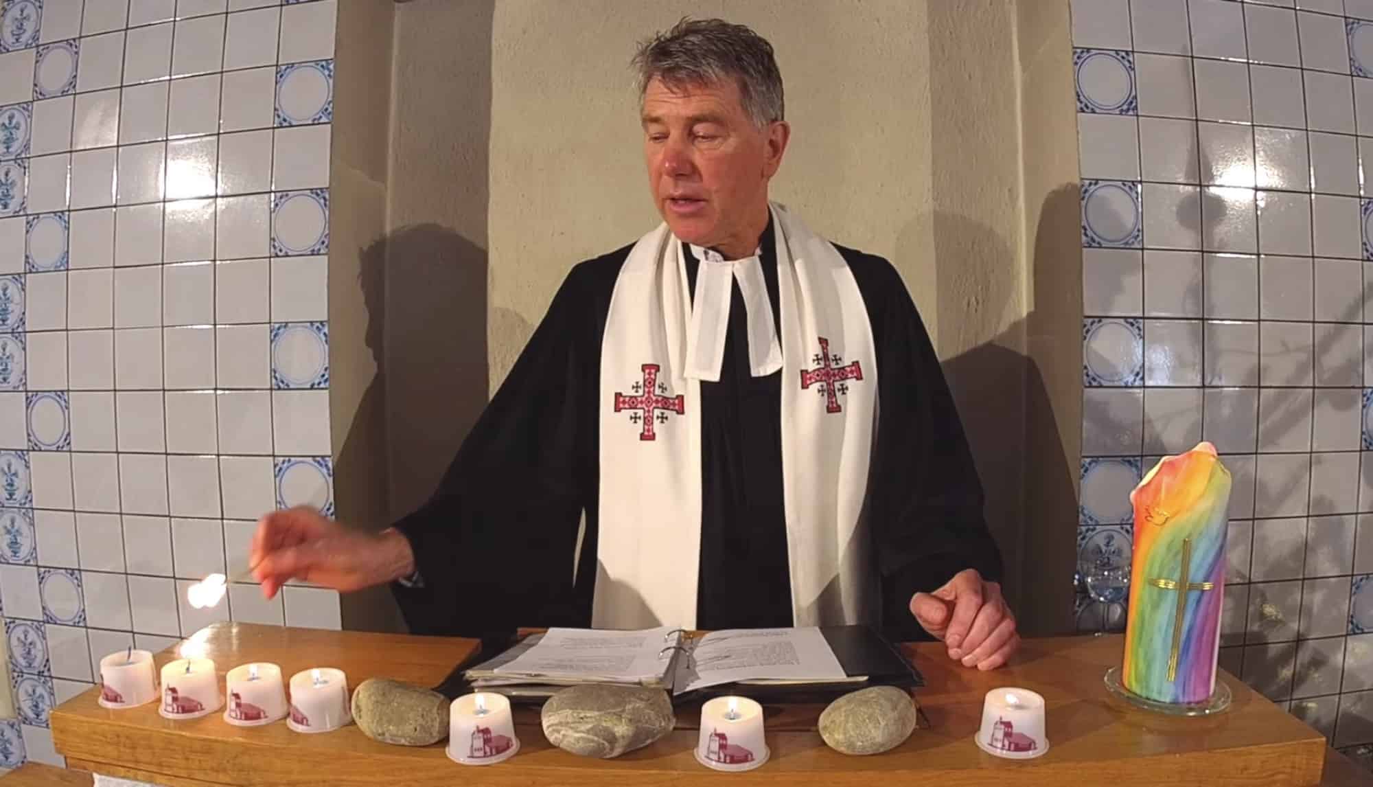 Pastor Rainer Chinnov auf der Kanzel beim Anzünden einer Kerze, die in einer Reihe mit Steinen und Kerzen aufgereiht ist.