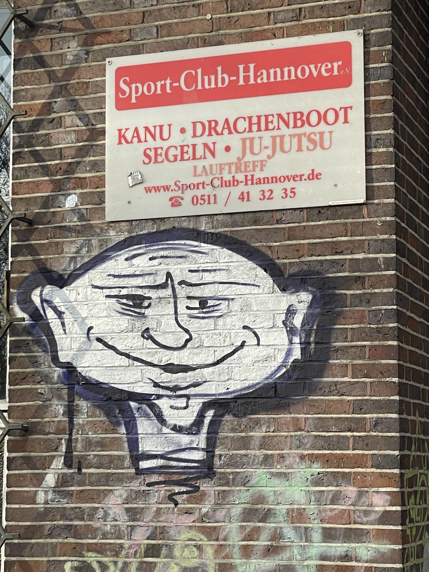 Schild des Sport-Club-Hannover an einer dunklen Ziegelstandwand mit asiatisch anmutenden Graffitikopf darunter.