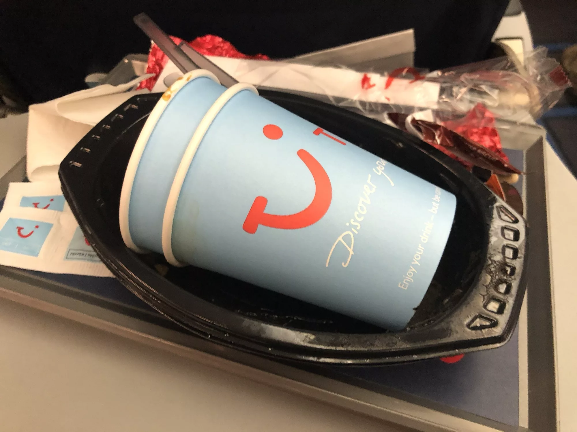 Hellblaue TUI-Kaffeebpappecher auf einem schwarzen Bordessentablett im Flieger.
