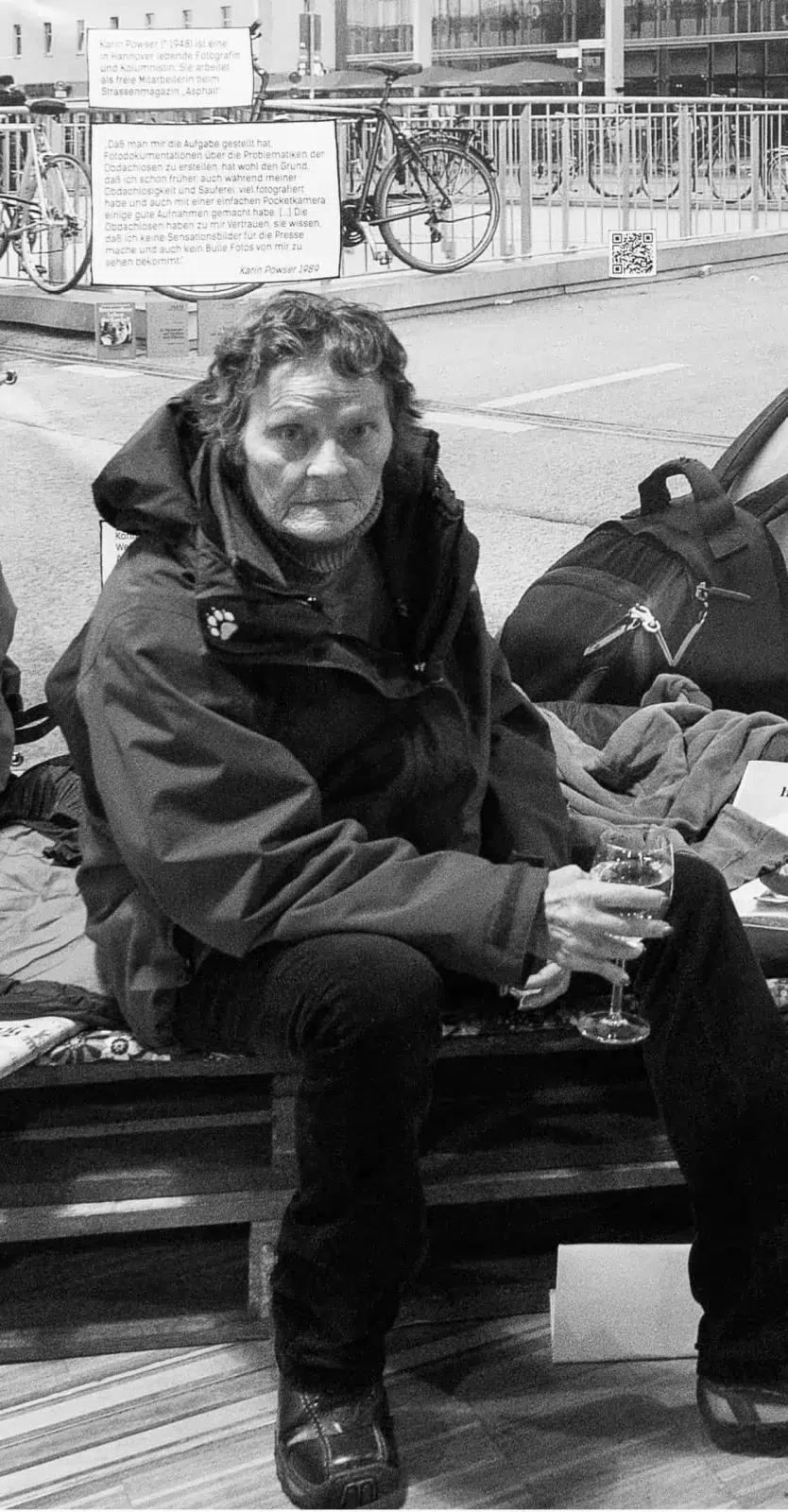Karin Powser sitzt auf Europaletten mit Schlafsack (scheinbar ein Obdachlosenschlaflager) im Fußgängerbereich in einer Innenstadt.