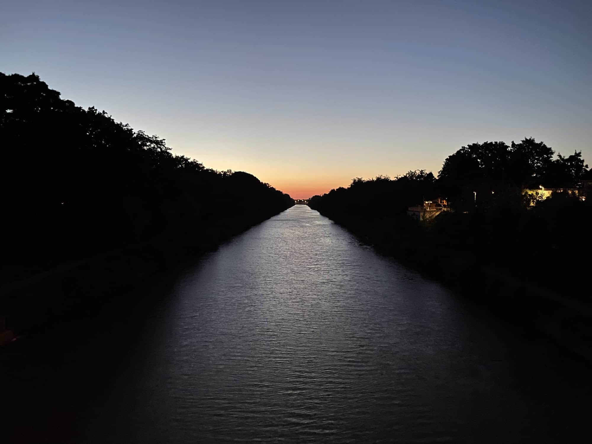 Blick von der Mittellandkanalbrücke Hannoversche Straße auf das Wasser und die gelb-orange Sonne am Horizont bei schwarzen seitlichen Kanalböschungen und hell-mittelblauen Himmel.