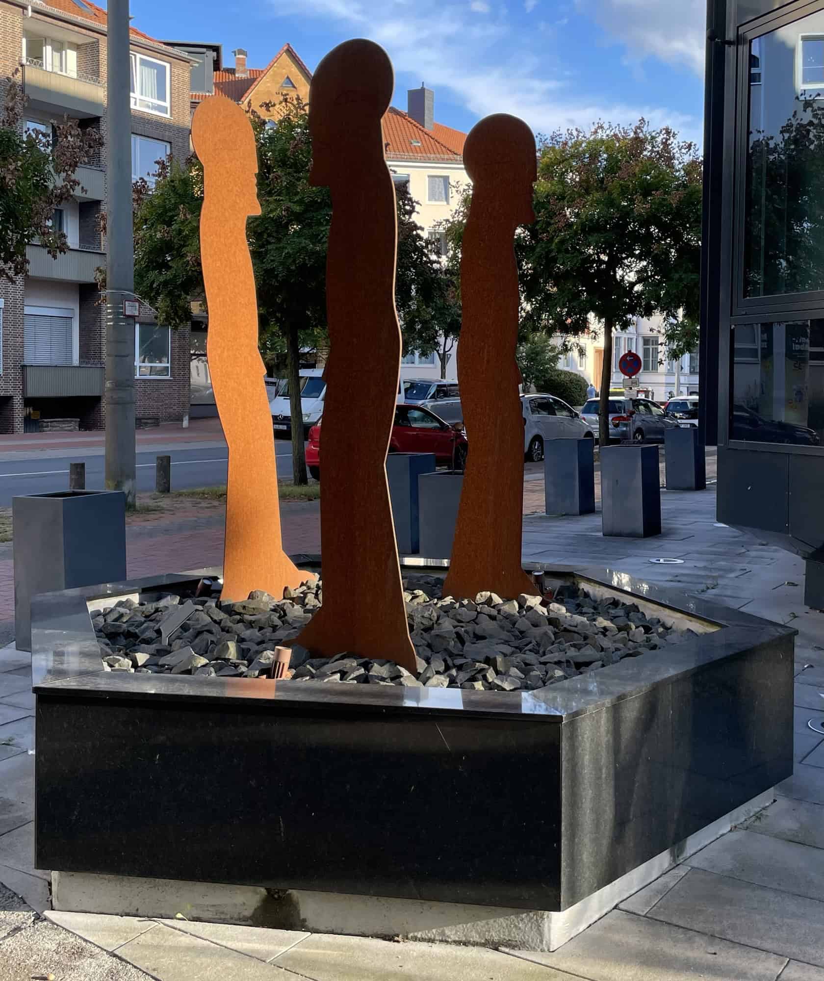 Kunst um Raum: Drei 2D-Figuren aus rostigem Metall in einem übergroßem Steinblumentopf.