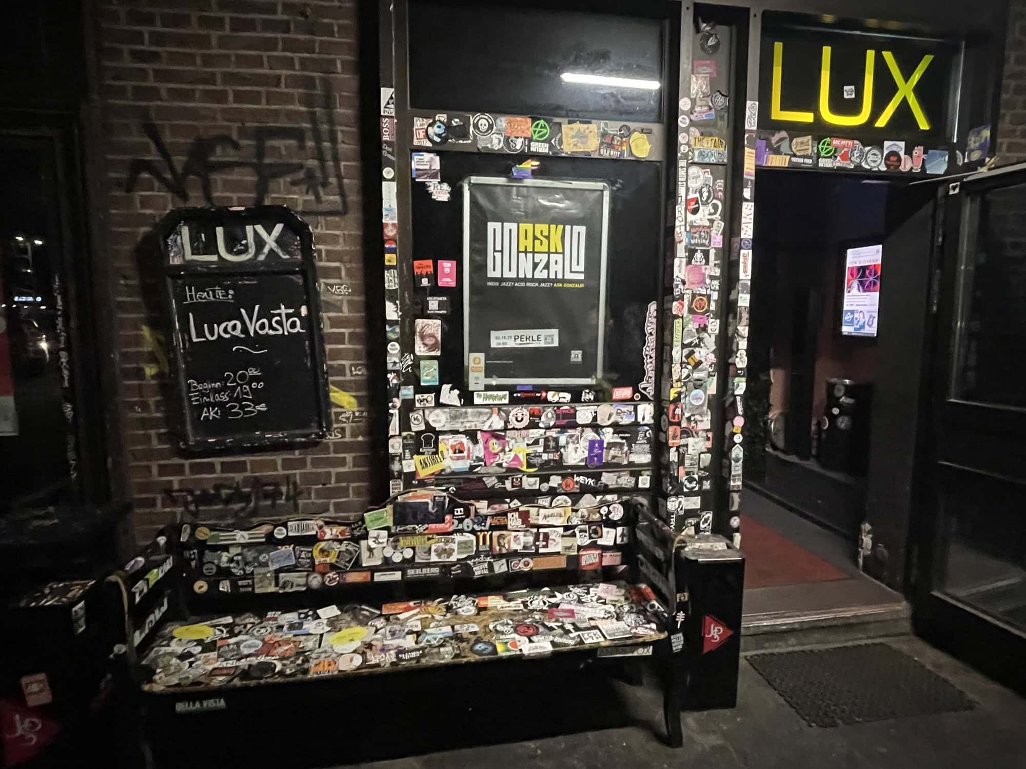 Der Einfang des Lux mit Tür und darüber dem gelben leucht-Lux-Schriftzug, links eine Bsnk. Alles viel mit Aufklebern beklebt.
