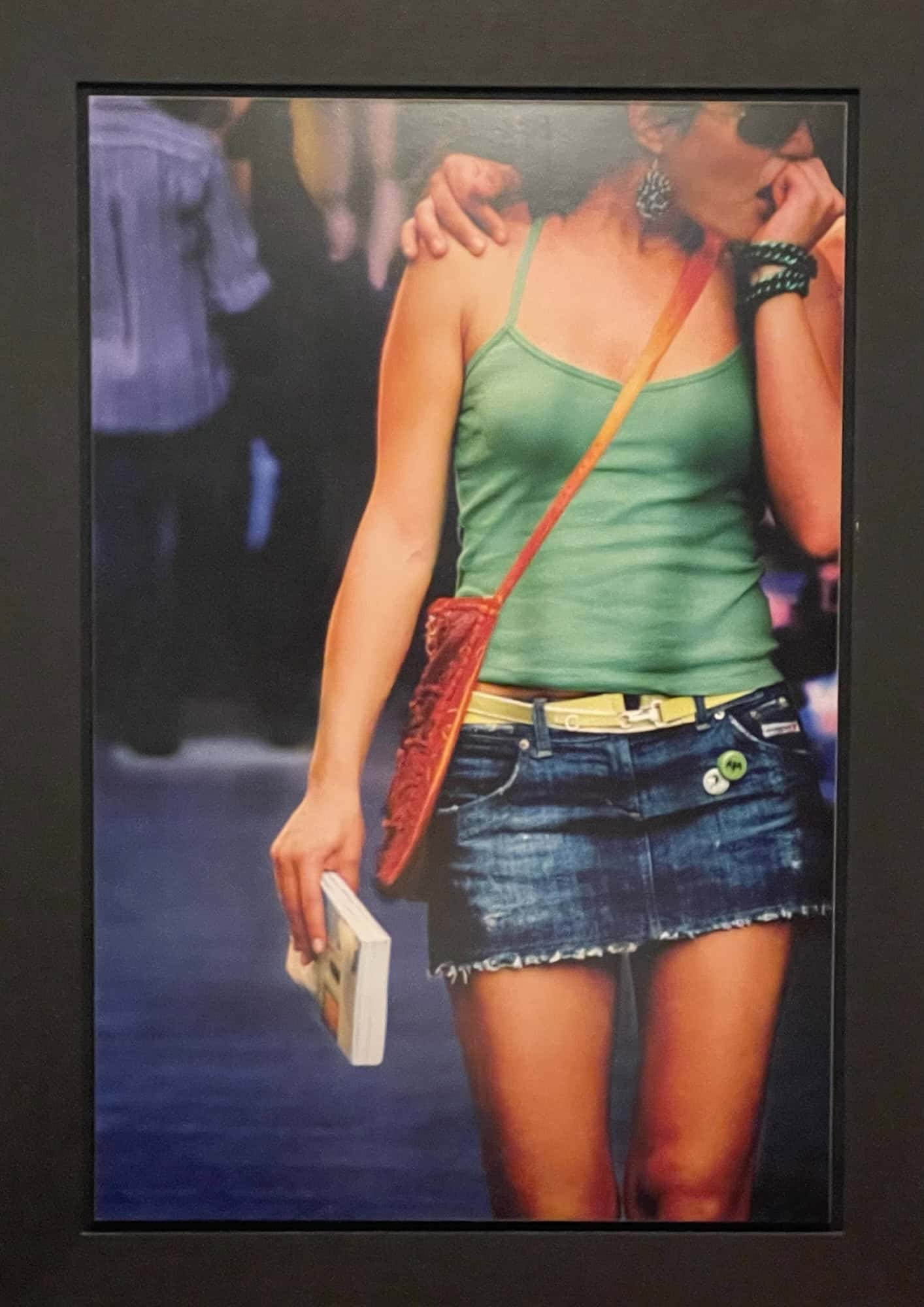 Portrait junge Frau mit blauem Jeansminirock und grünem Spaghrttiträgertop, Mann hat Arm auf ihrer rechten Schulter, sie trägt runde-schwarze Sonnenbrille, ihr Kopf ist obe abgeschnitten.