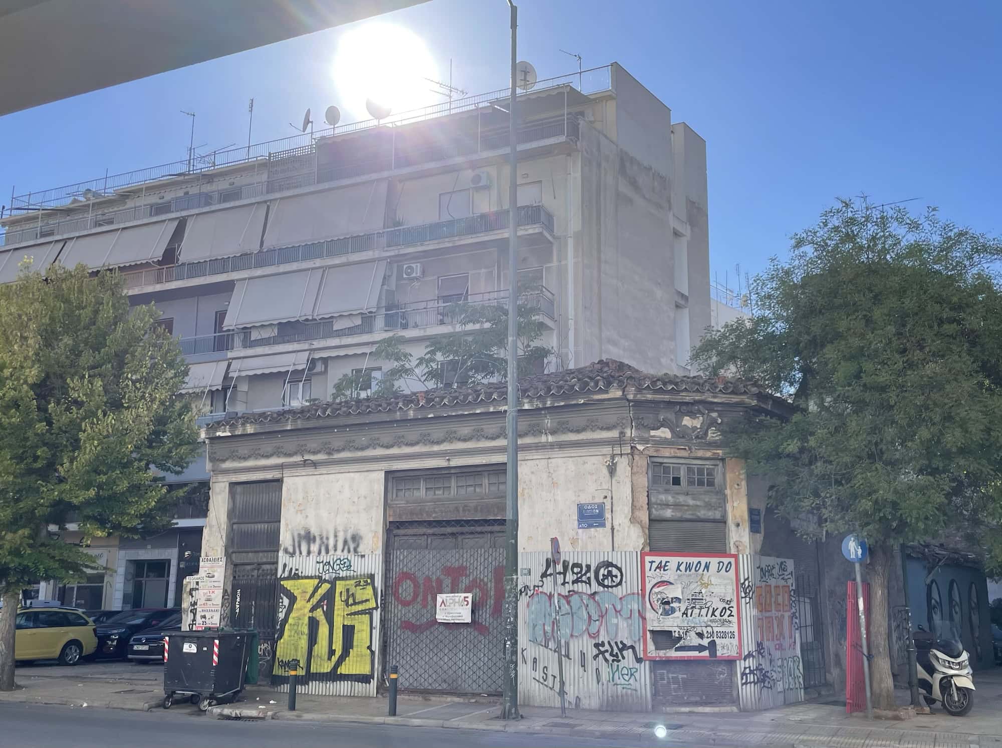 Kleinesres Haus mit GeFfitis und Reklame, dahinter ein Wohnblock über dem die Sonne hell steht.