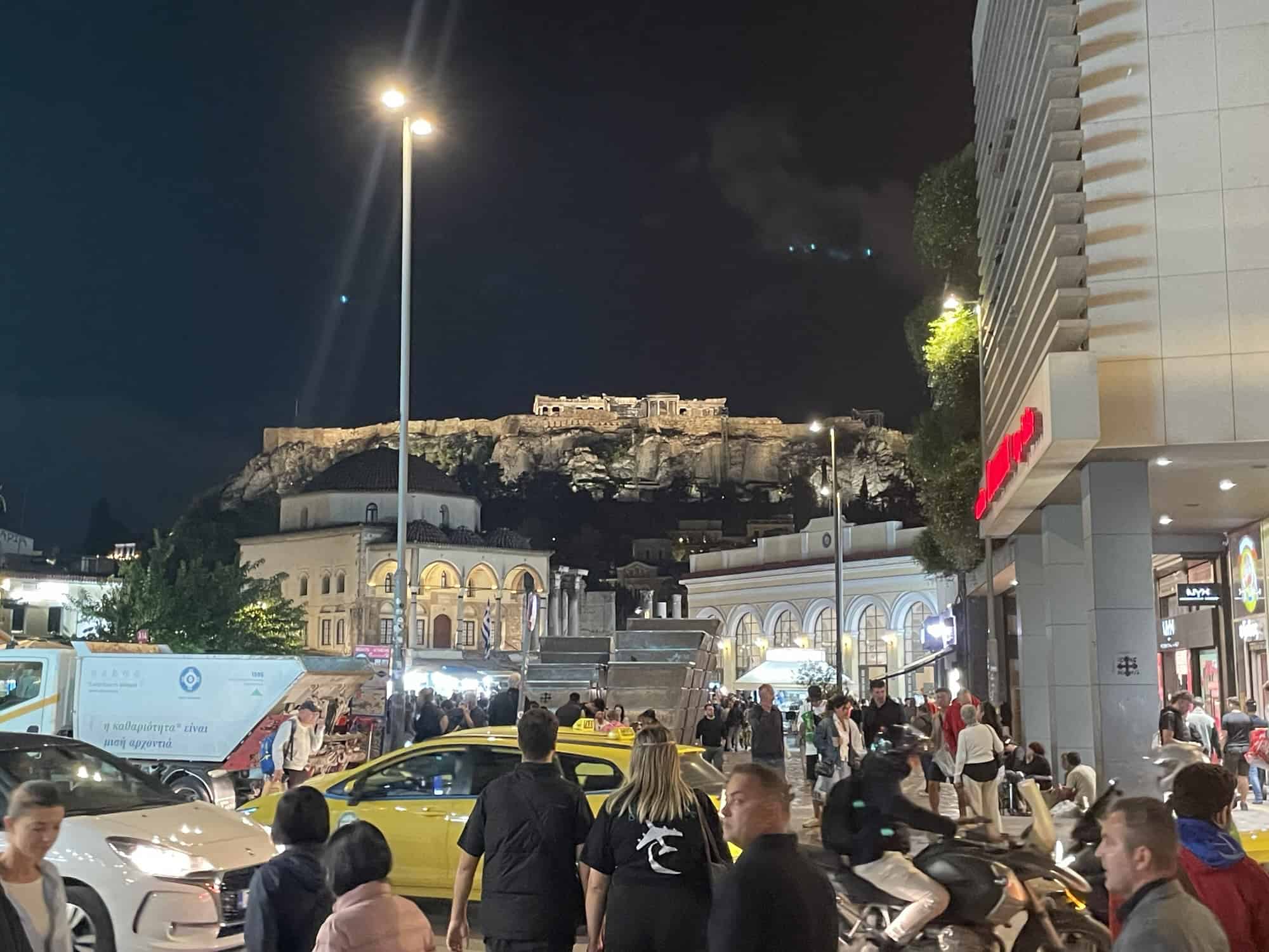 Gewimmel auf der Straße im Dunkeln, im Hintergrund thront die Akropolis.