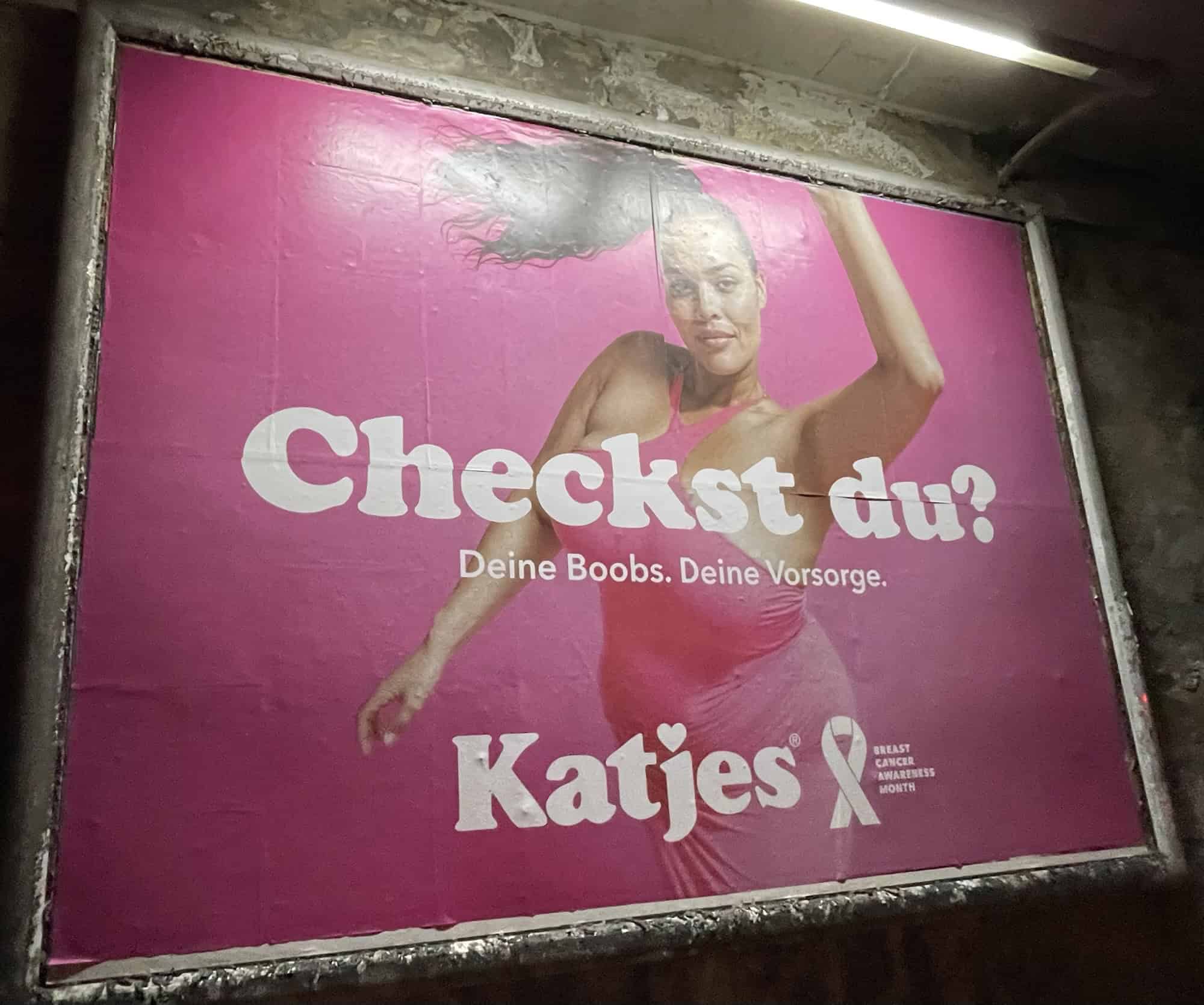 Außenwerbeplakat 'Checkst du!' rosa von Kajes mit abgebildetet Frau zum Brustkrebsmonat Oktober, dunkelhäutige Frau mit fliegendem Zopf zur Seite, linke Hand hochgereckt und in somnerluchen lässigem Rosakleid.