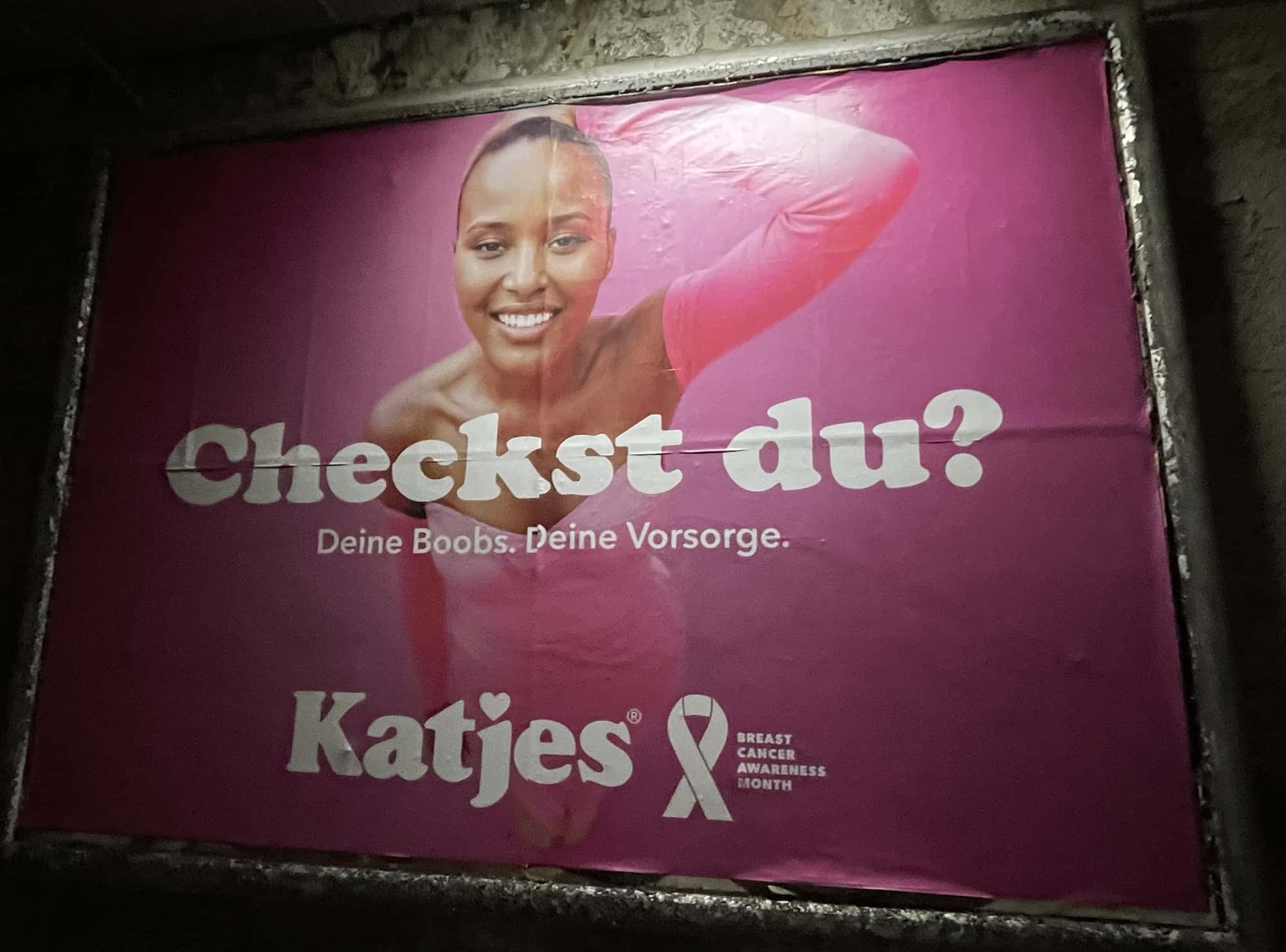 Außenwerbeplakat 'Checkst du!' rosa von Kajes mit abgebildetet dunkelhäutige Frau zum Brustkrebsmonat Oktober, gleiche Frauenabbildung wie vorangegangens Bild.