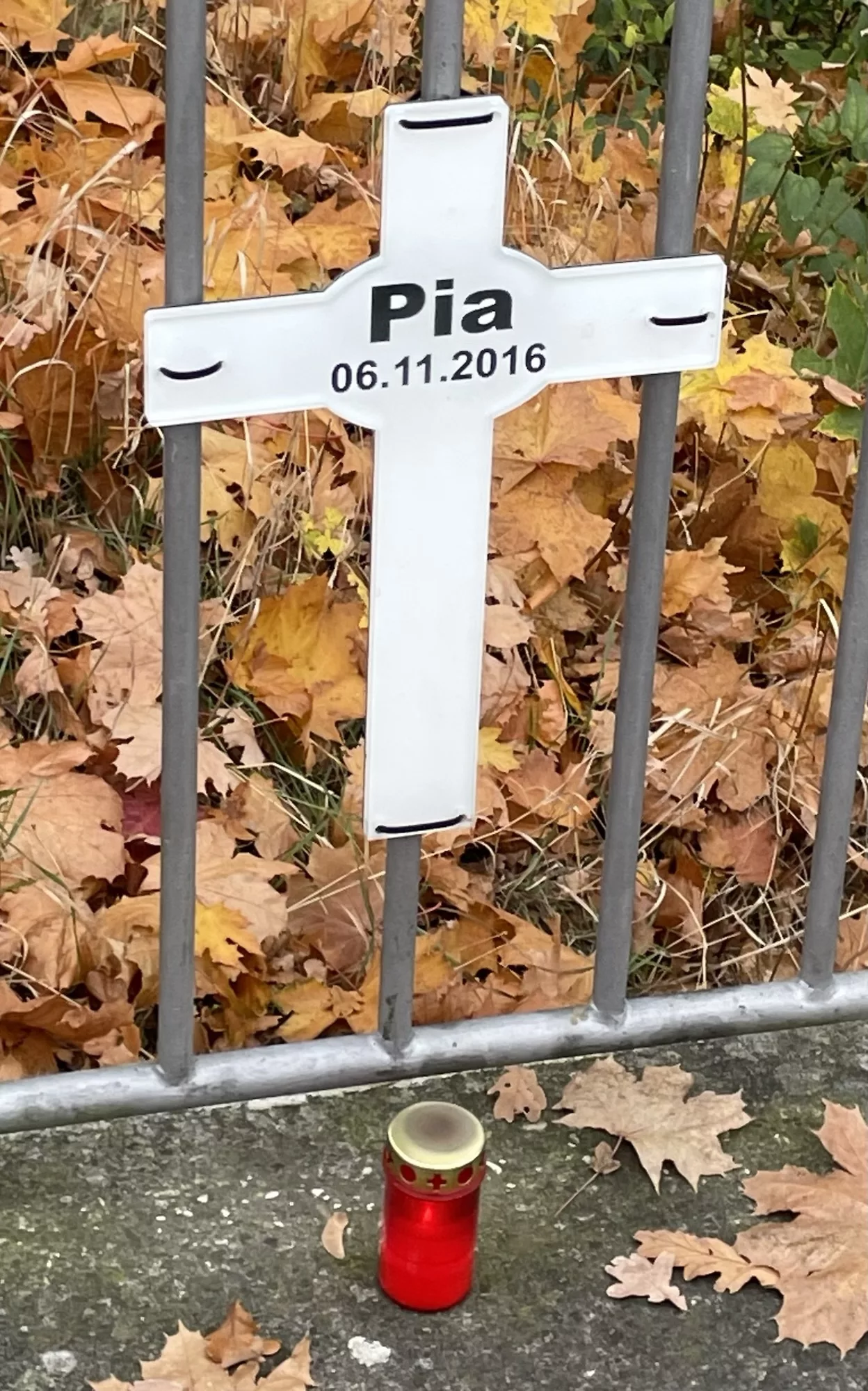 Weißes Plastikkreuz on Pia an Metallzaun des Mittellandkanals mit btennenfem Totenlicht davor.