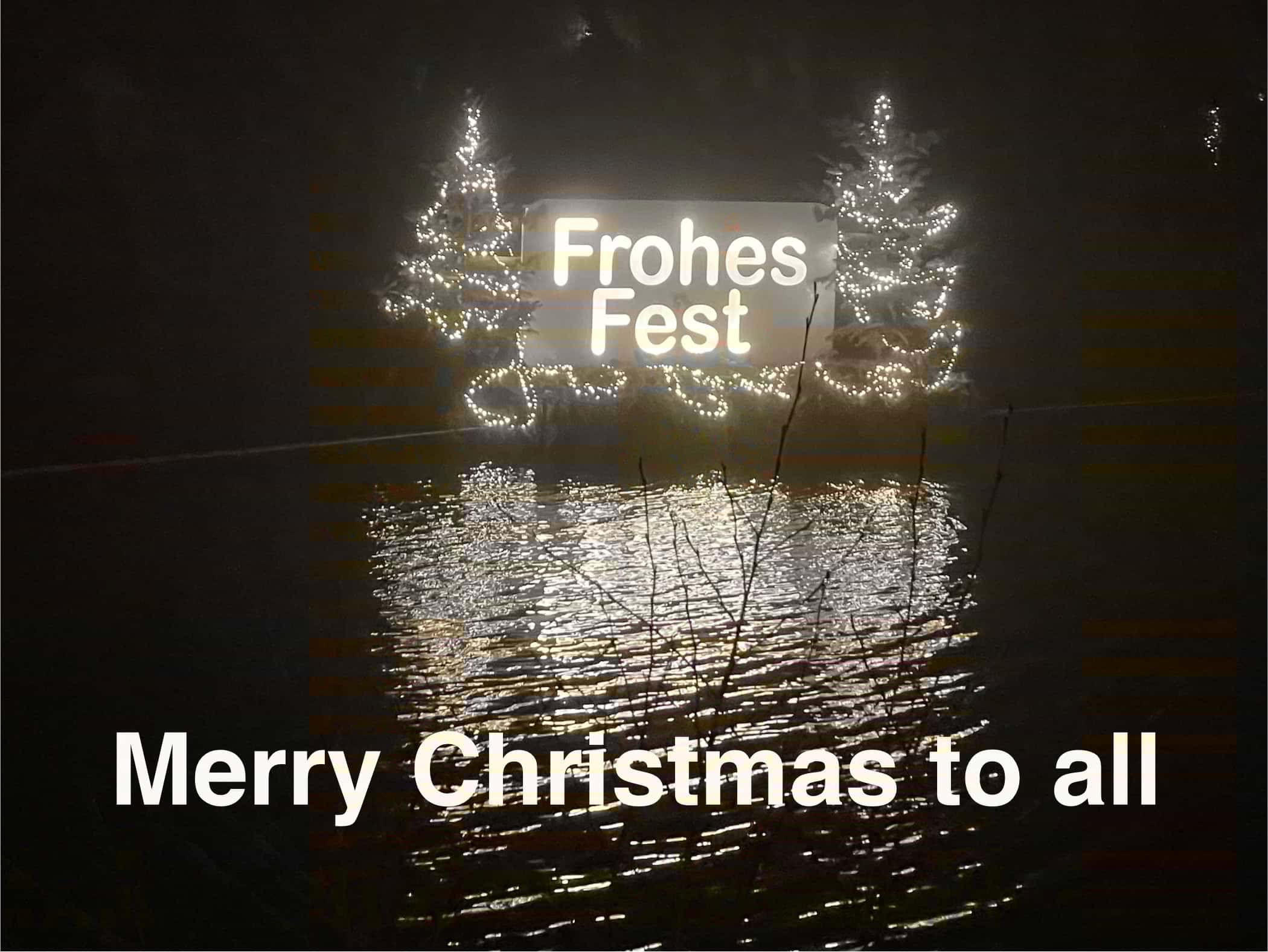 Nächtlicher Teich mit weiß leuchtendem 'Frohes Fest'Tafel zwischen erleuchteten Tannen, unten im Bild ist der Schriftzug 'Merry Christmas to all'. 