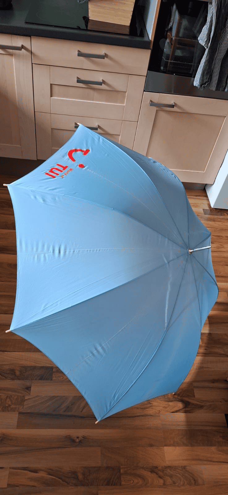 Hellblauer aufgespannter Regenschirm mit rotem TUI-Schriftzug und Smily auf dem Holzfußboden vor hellem Holzschrank.