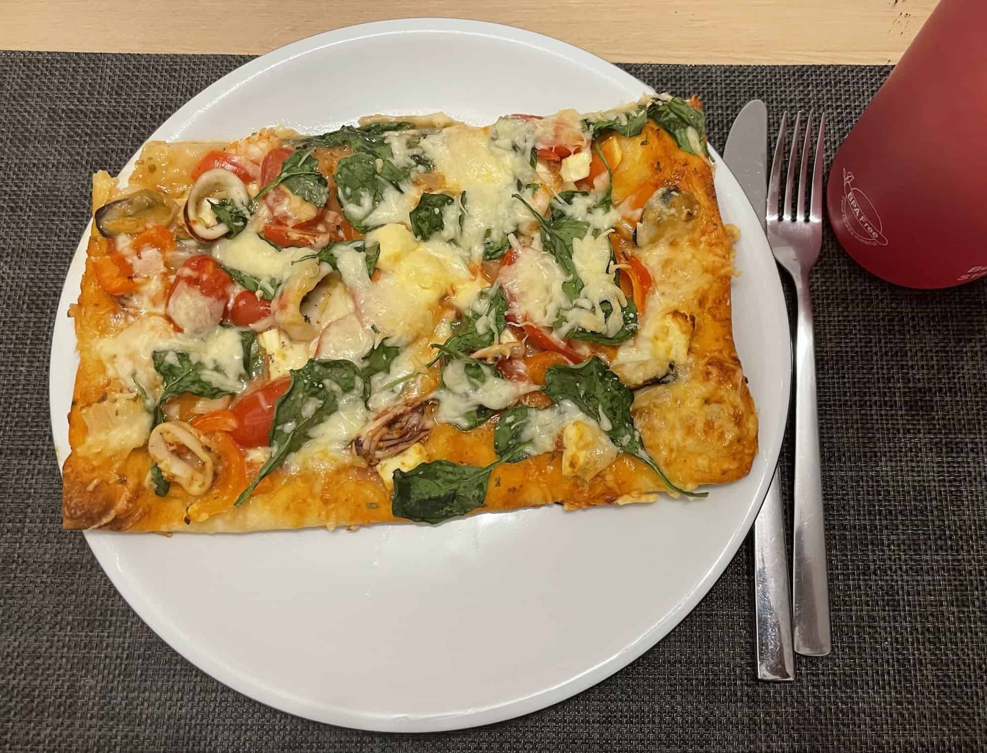 Eßteölrr mit großem Stück Meeresfrüchtepizza, daneben Besteck und magentafarbene Trinkflasche auf schwarzem Set.