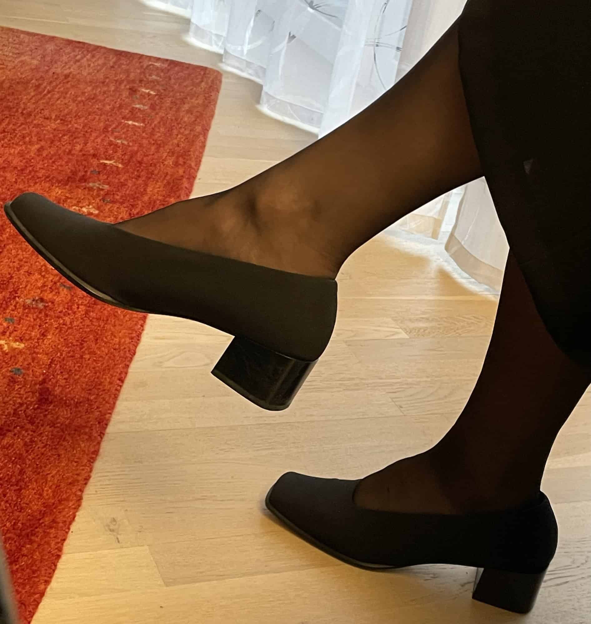 Frsuenbeine mit schwarzen Pumps und schwarzer recht blickfichter Strumpfhose sitzend auf Parkett und Teppich.