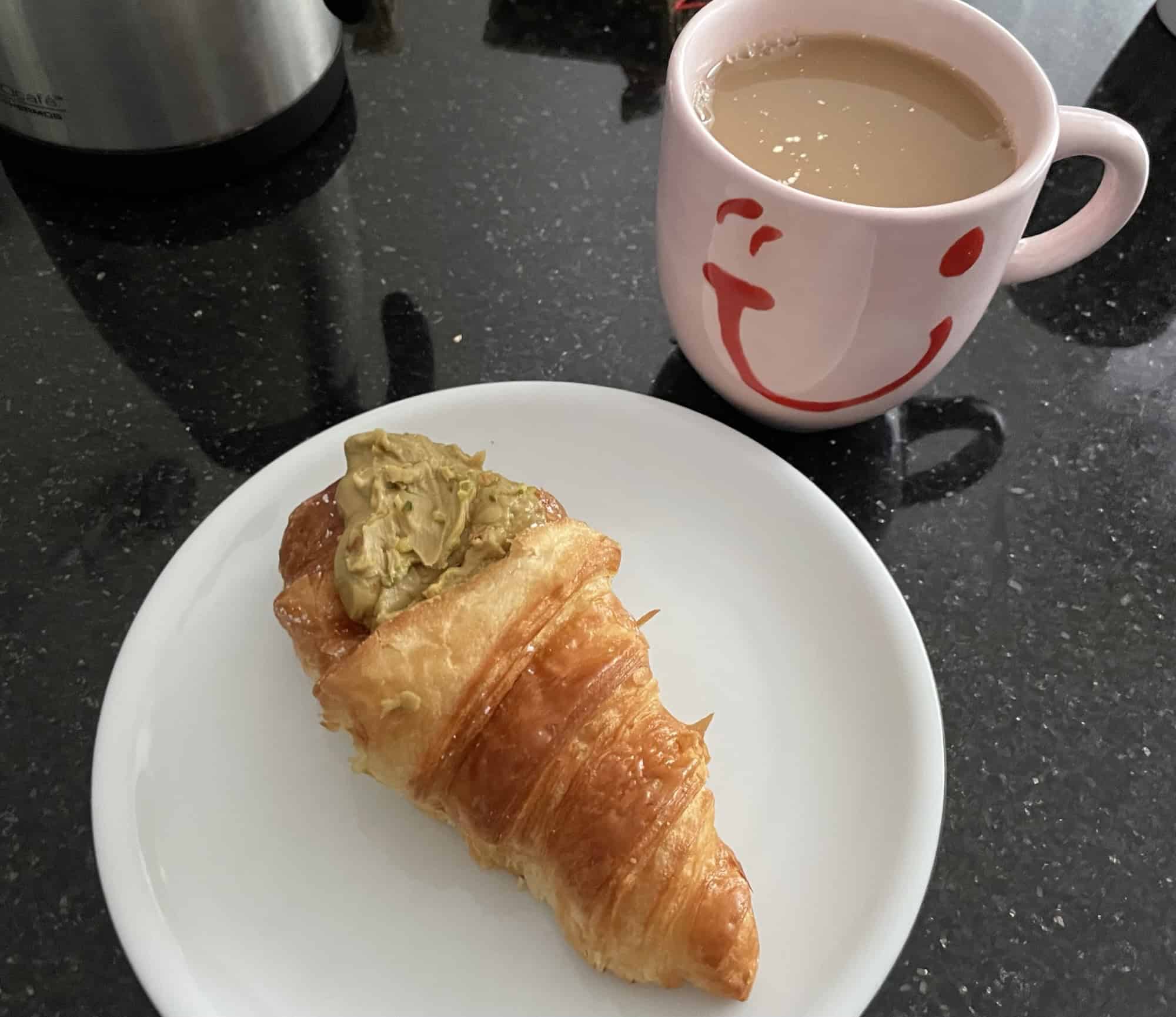 Ein Cornetti al Pistacchio auf weißem Teller, Metallkaffeekanne und rosa Becher mit rotem Urlaubdlächeln darum.