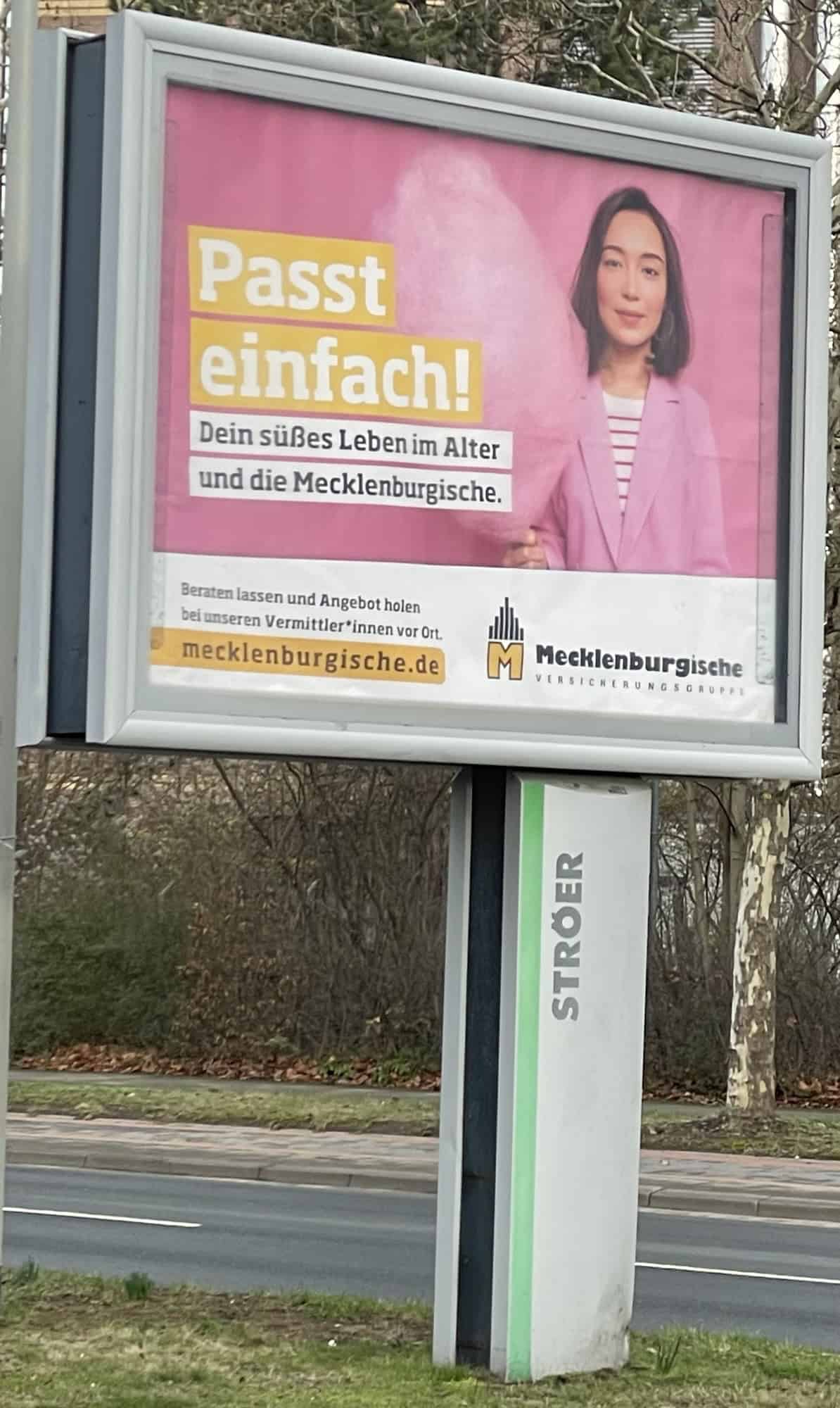 Grosßwerbubg auf Mittelgrünstreifen der Allee mit Werbung der Mecklenburger Versicherungen mit Frau in rosa und rosa Zuckerwatte und Slogan: Passt einfach! Dein süßes Leben im Alter und die Mecklenburgische.