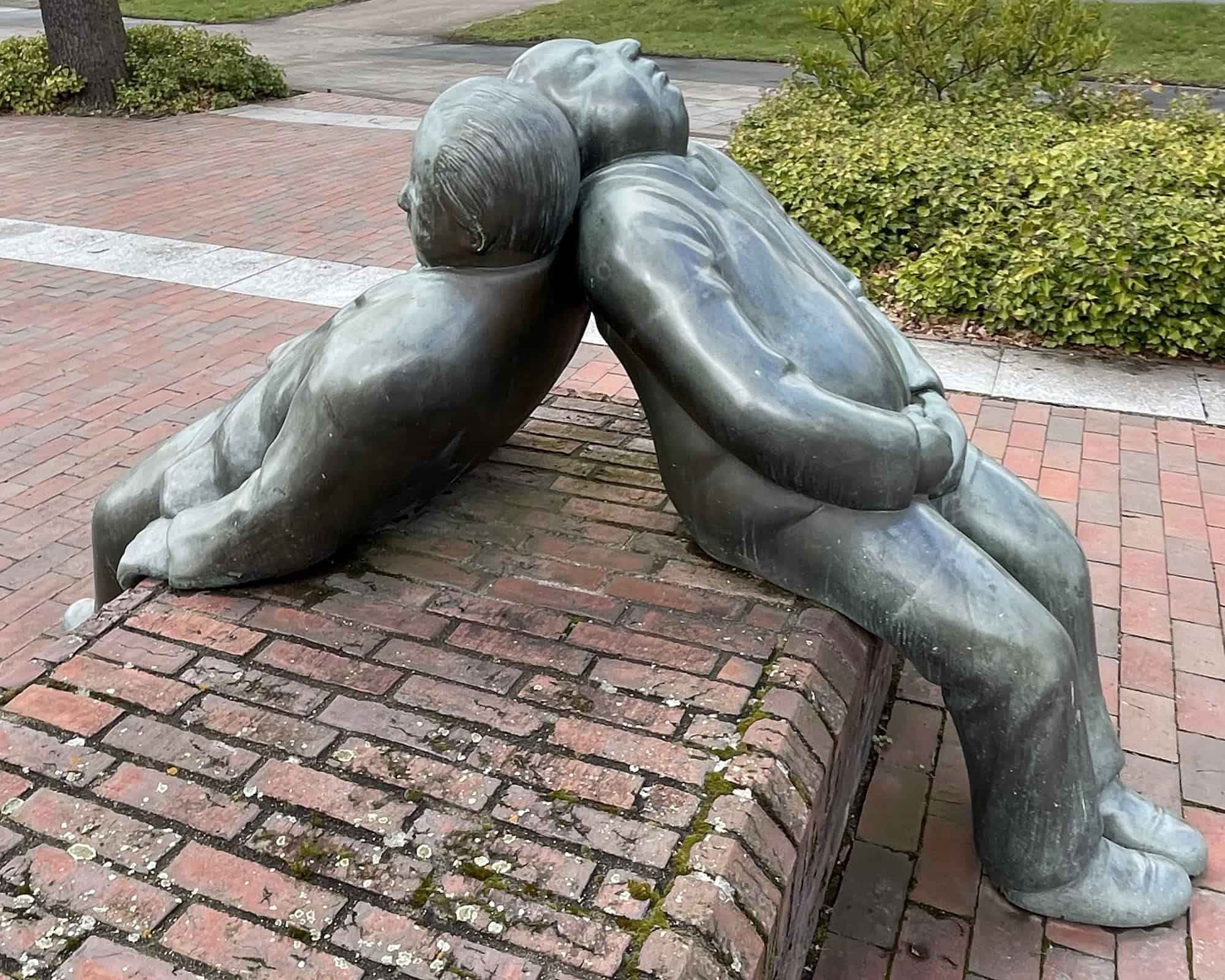 Zwei sich Rücken and Rücken anlehnende, schlafende und sitzende männliche Metalkskulpturen.