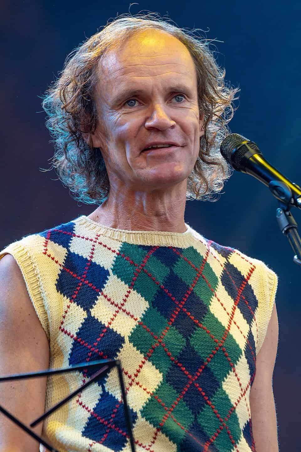 Portrait von Olaf Schubert mit markanten buntem Rautenpullunder in gelb auf der Bühne mit Mikro und Notenständer.