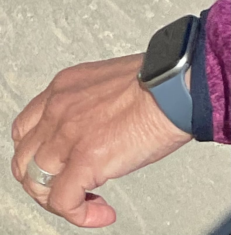 Braune Hand mit Apple Watch mit blauem Armband, Ring am Ringfinger und lila Fleecejacke.