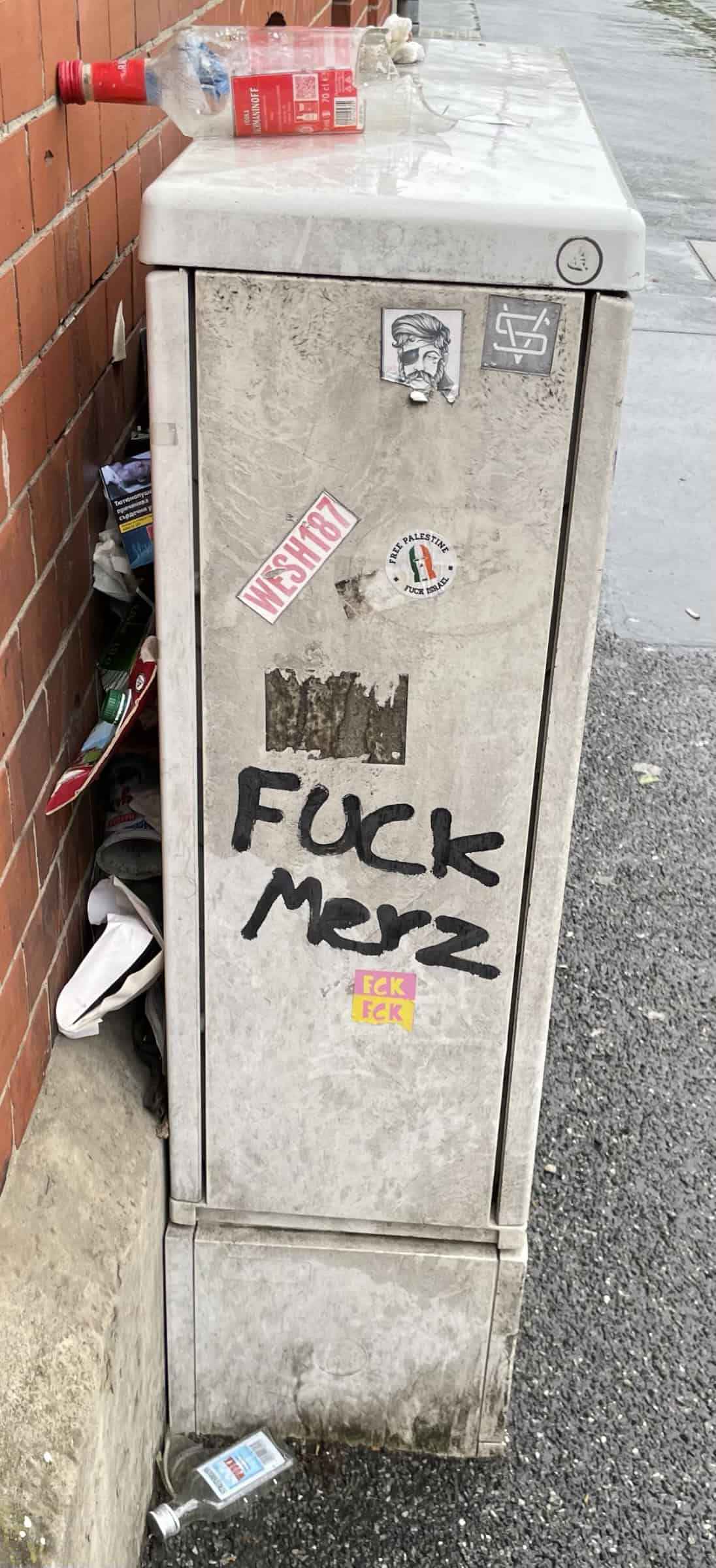 Grauer Elektrokasten an Wand mit Müll, keeren Flaschen und Tagging: Fuck Merz