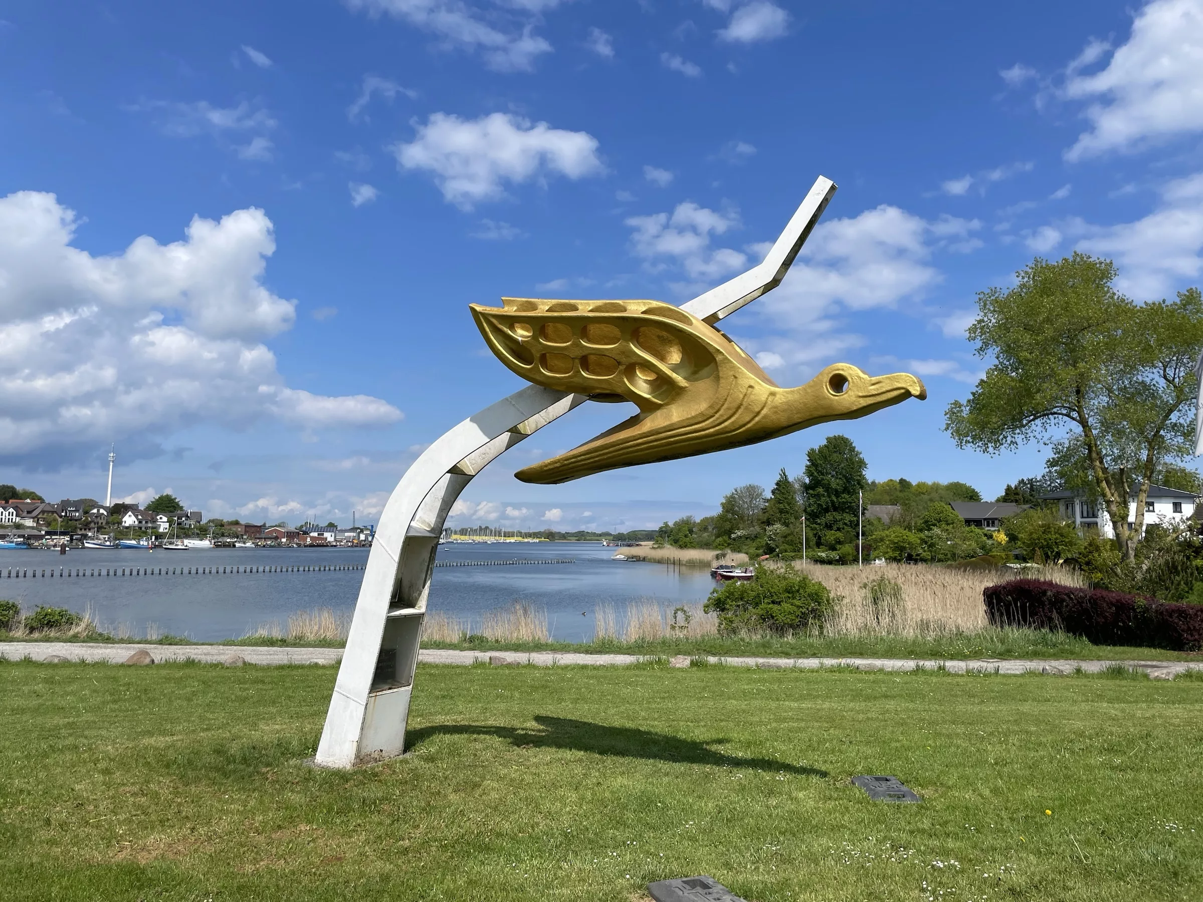 Kunstskulptur übergroßer goldener Metalvogel an weißer schräger Stange.