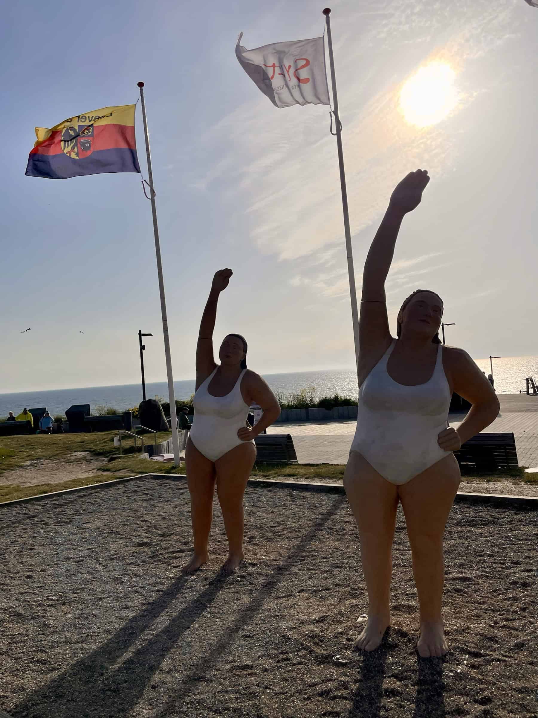 Zwei Frauenskulpturen im Badeanzug mit Gymnastikbewegung, dahinter zwei Fanen im Wind, die Promenade und der Strand von Wenningstedt.