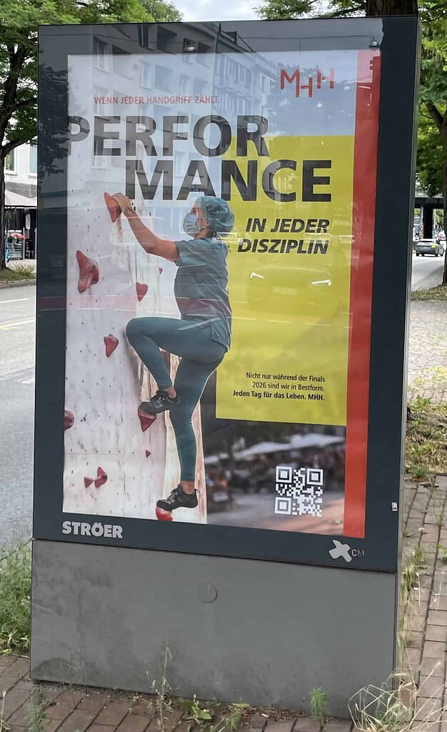 Straßenreklame Tafel in klein mit Werbung der MHH: Person in grüner Funktionskleidung mit Mundschutz und Op-Haube klettert eine Indoor-Kletterwand hoch, dazu Slogen: WENN JEDER HANDGRIFF ZAHLT. PERFORMANCE IN JEDER DISZIPLIN.