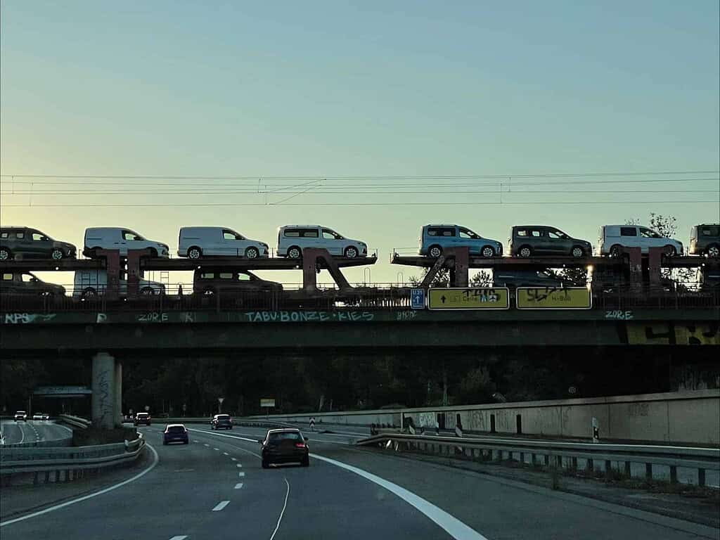 Brücke über Schnellstraße im Abendlicht, auf der ein Güterzug mit zweistöckig geladenen Caddy-Autos steht.