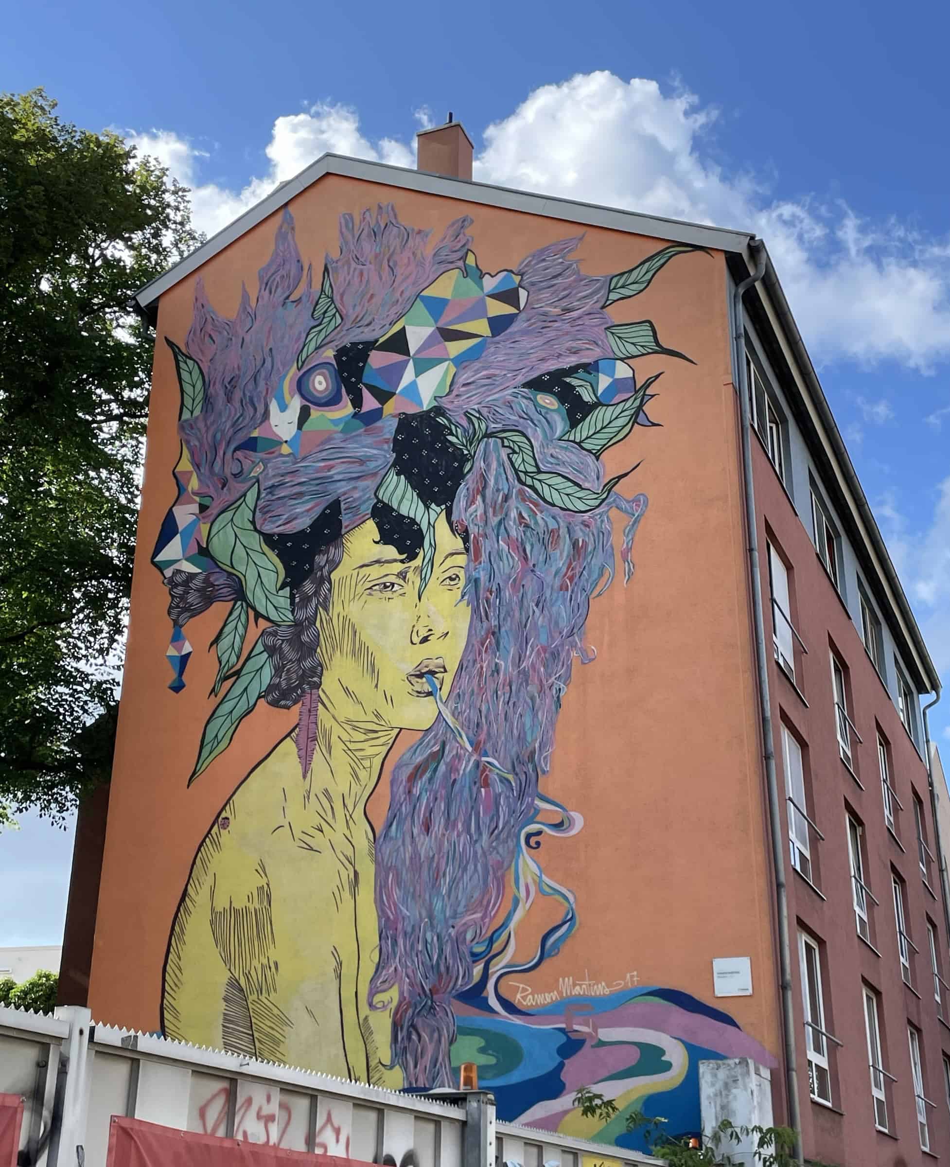 Großwandbild an Hauswand mit Portrai einer Frau (Oberkörper) mit schwarzen Haaren und einem oppulenten Hut/Haarschmuck mit lila runterhändem Haarzopf.