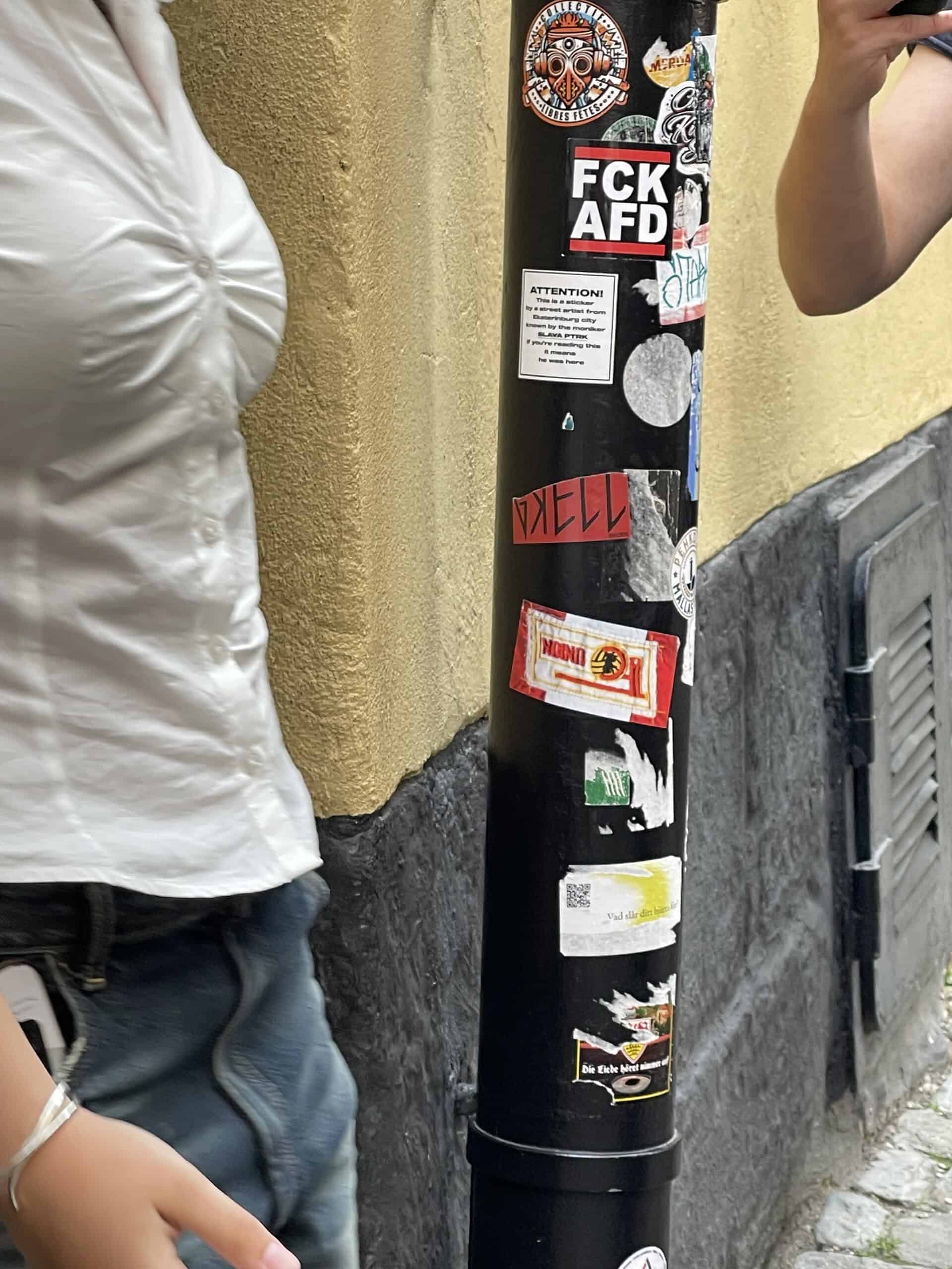 Dieser schwarze AfD-Aufkleber an Regenwasserfallrohr, Frau von links tritt ins Bild, rechts ein Arm einer anderen Person.