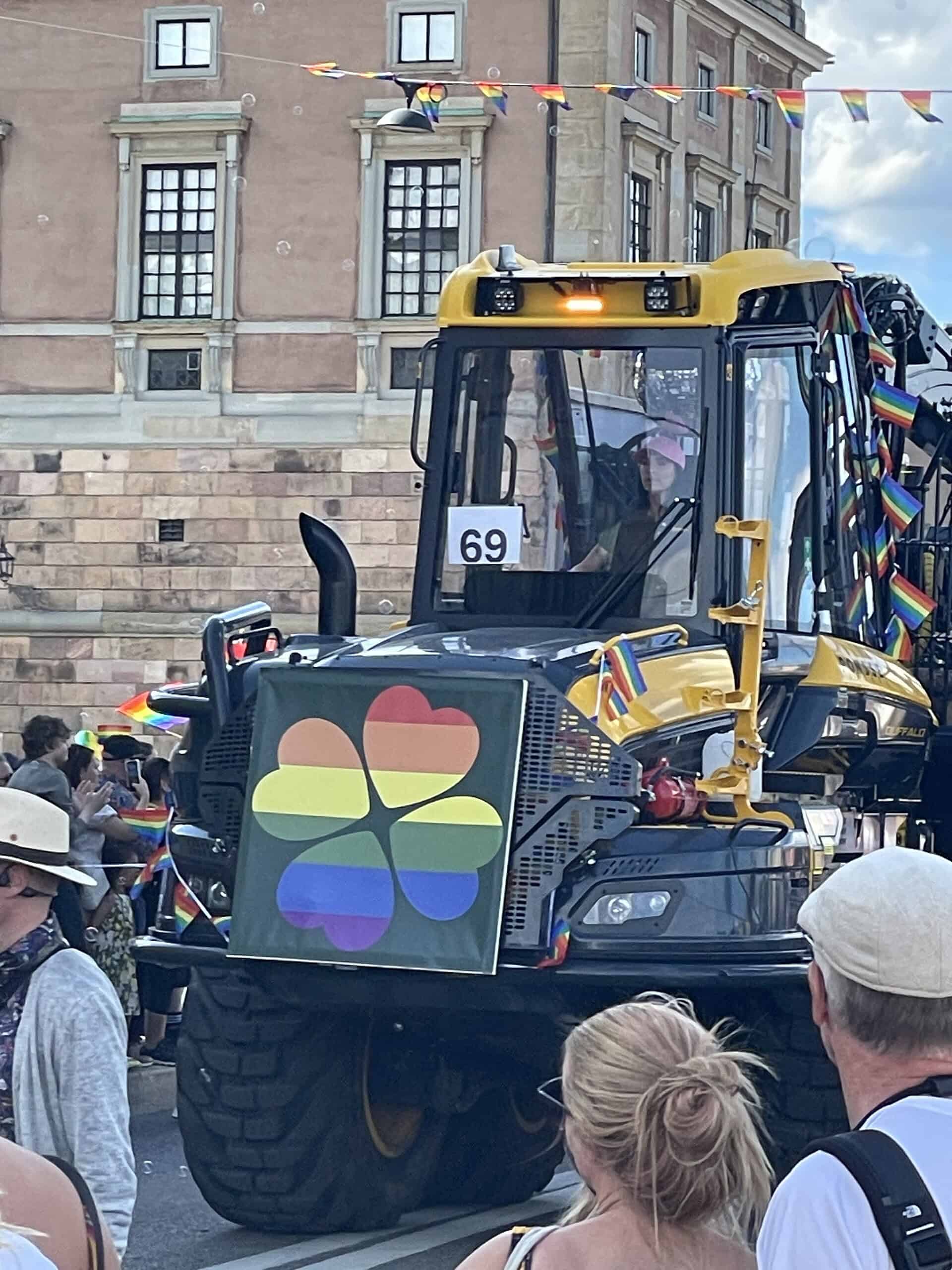 Gelbes Baufahrzug, gefahren von einer Frau mit rosa Arbeitshelm, vorne am Truck vier Kleebletter in Pride-Farben.