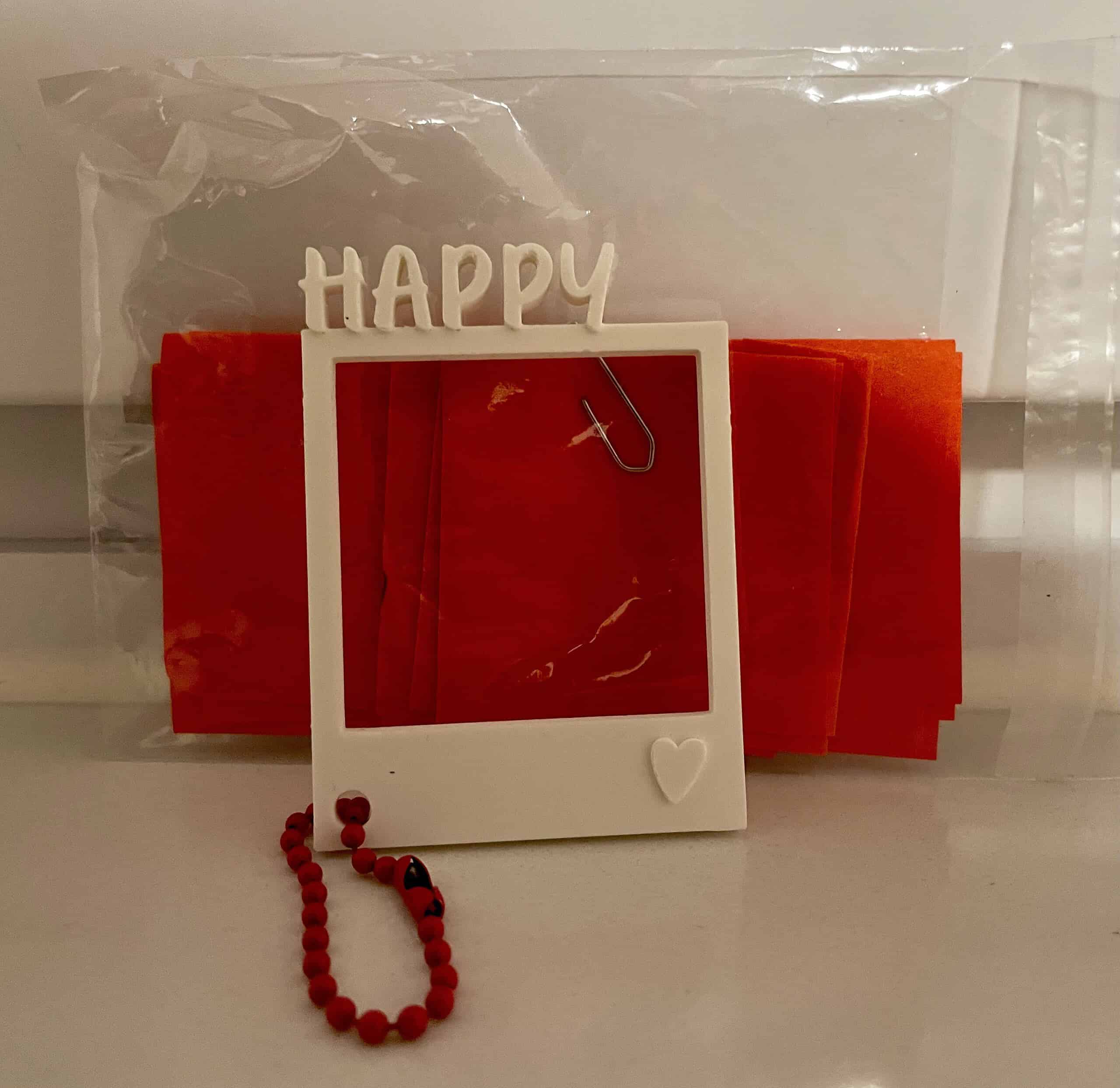 Durchsichtige Tüte mit roten kleinen Zetteln, davor ein kleiner weißer Bilderrahmen ohne Bild mit 'Happy'-Schriftzug oben und rote Kette unten.