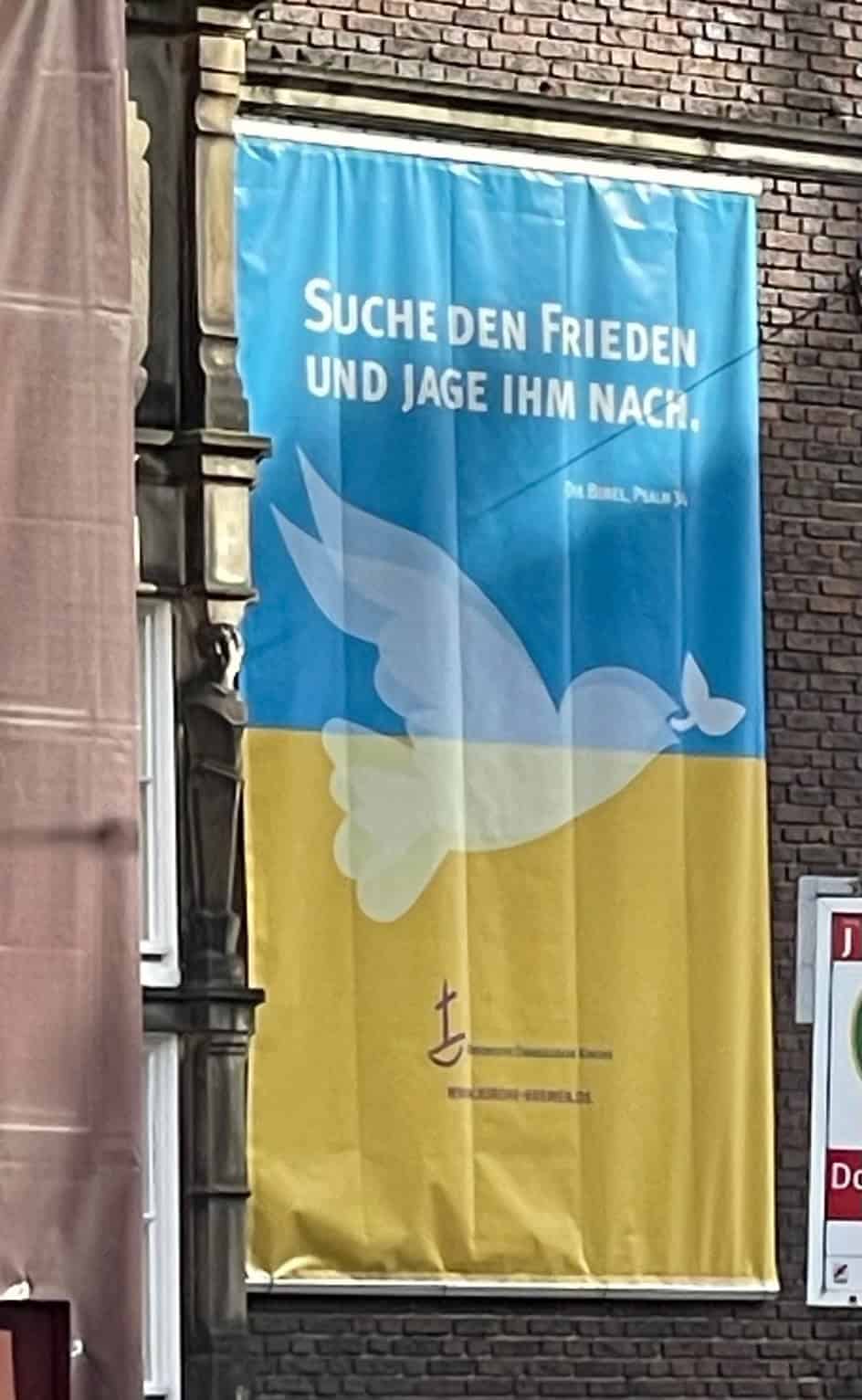 Großes gelb-blaues Plakat: 'Suche den Frieden und jage nach ihm' mit Friedenstaube.